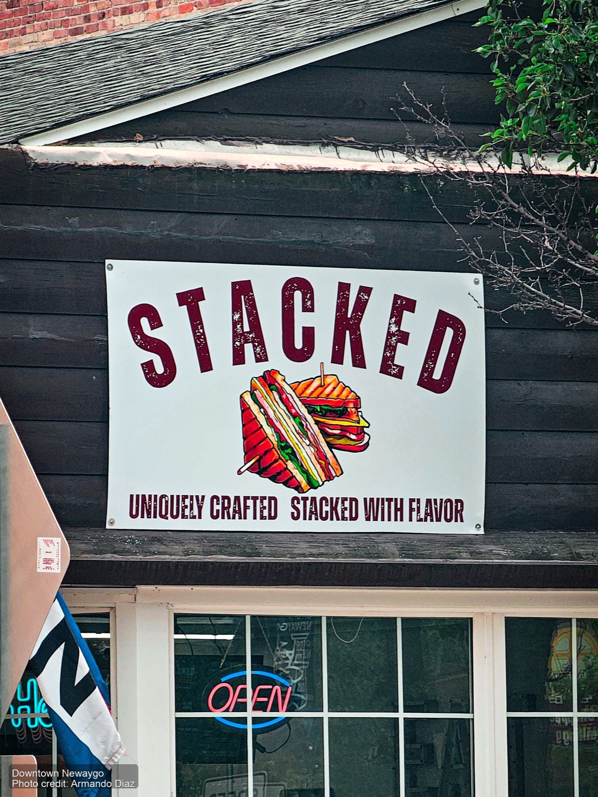 Stacked sandwich shop sign on a black wooden storefront in downtown Newaygo, Michigan, with a neon Open sign in the window below